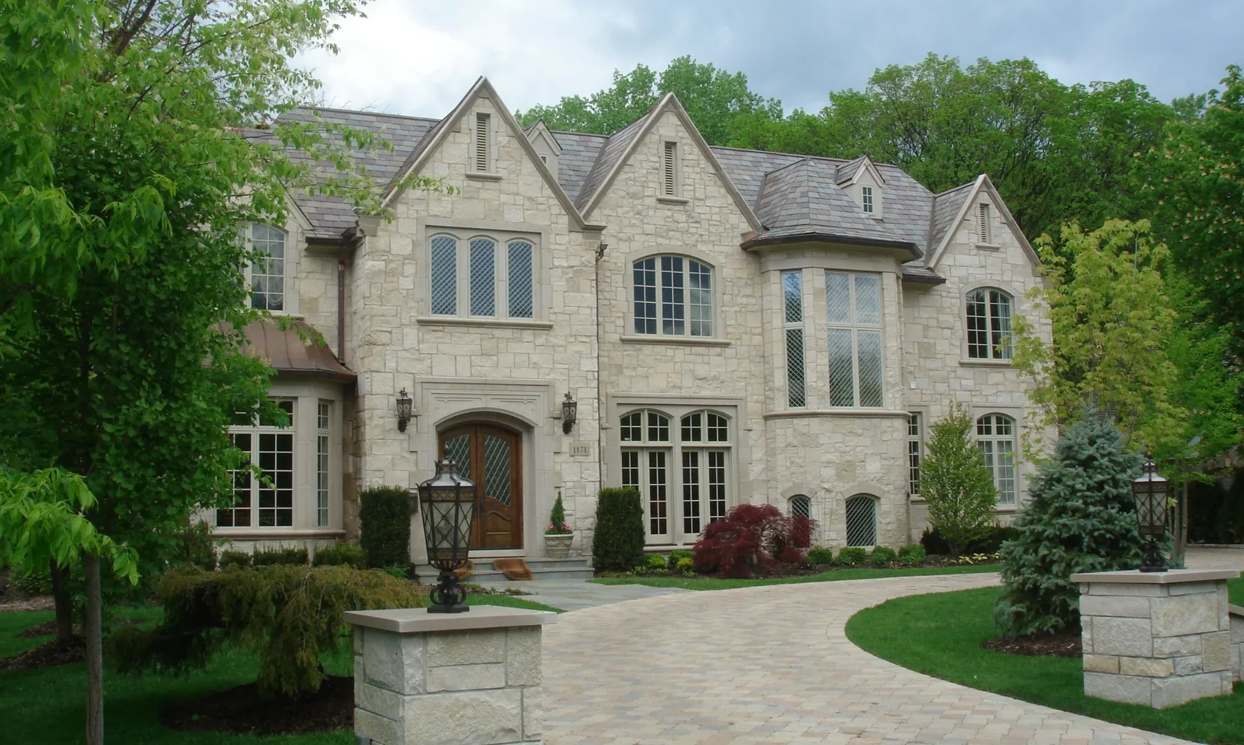 Light stone residence with multiple gables, slate roof, curved drive and mature landscaping.